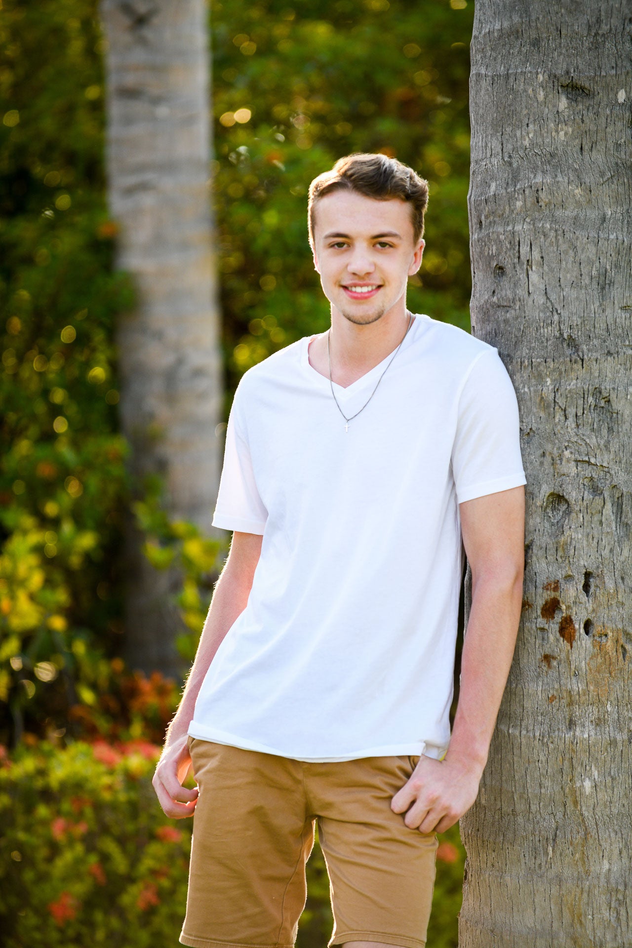 Man in a white shirt and beige pants leaning against a tree in a park setting. Cayman Islands senior portrait photographer