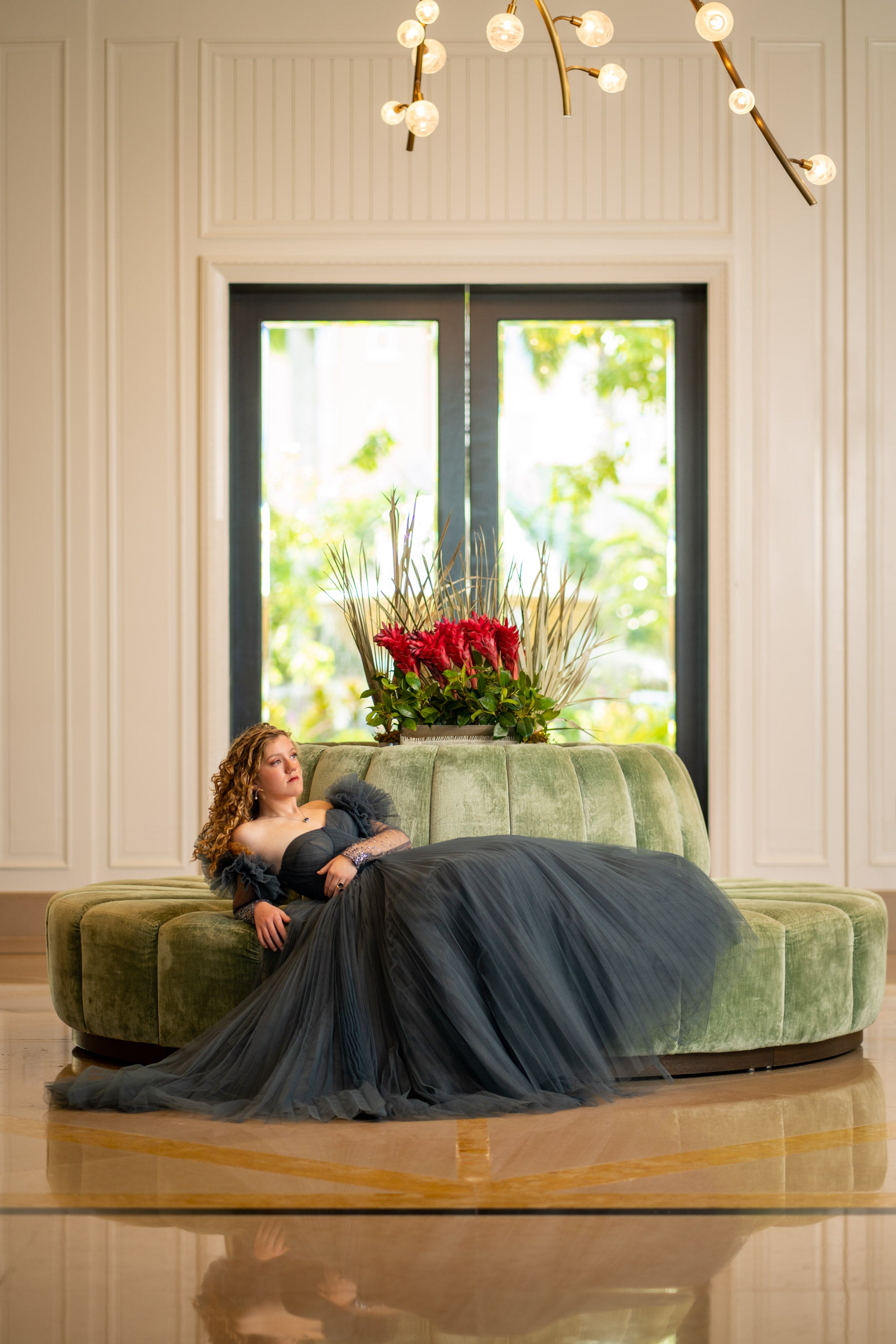 Woman in a long black dress sitting on a green couch with flowers in a room with large windows. Senior portraits Ritz-Carlton Cayman Islands 