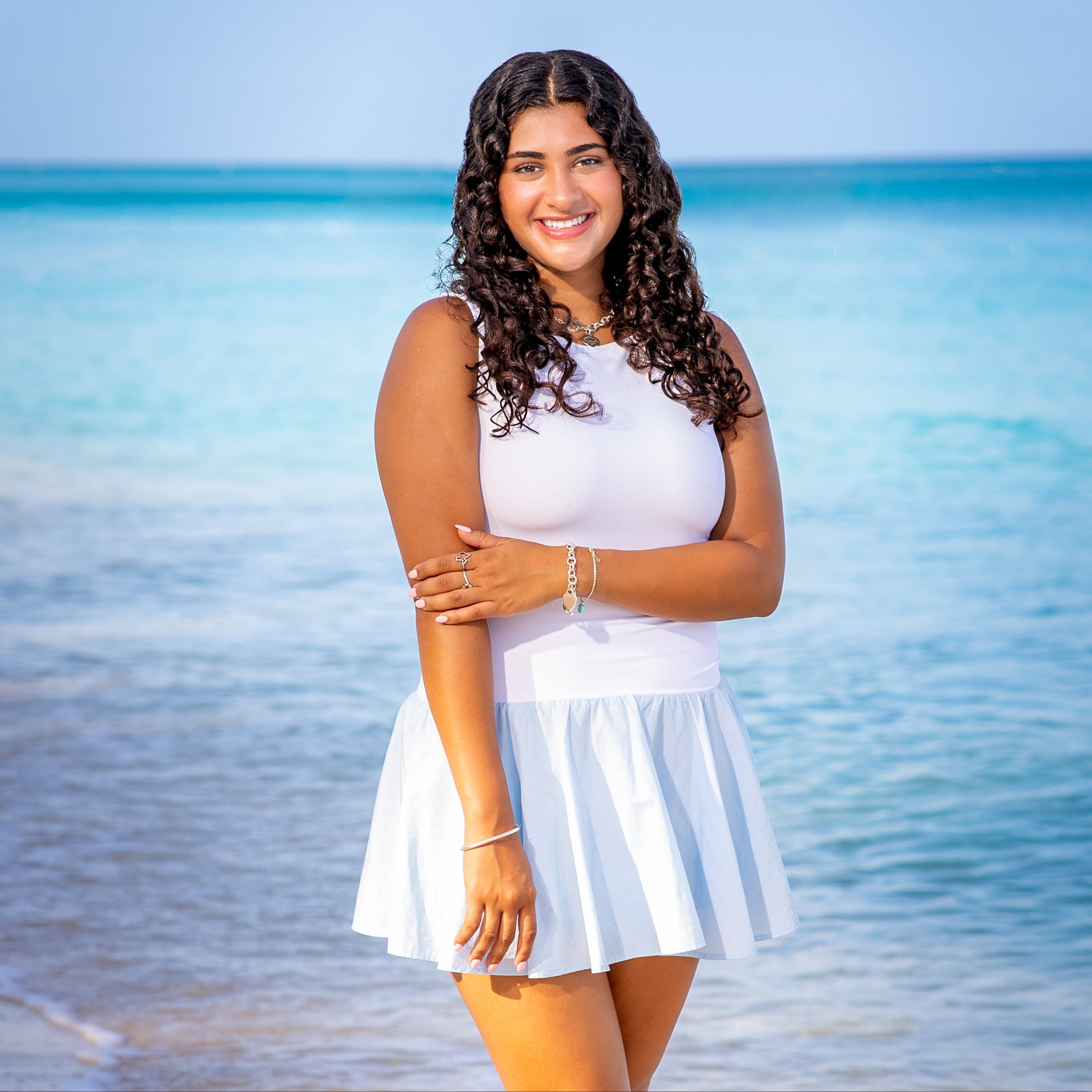 Woman in a white dress standing on a beach with clear blue water and sky. Cayman Islands senior portraits