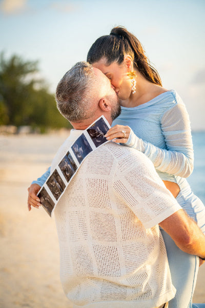 Man holding ultrasound images while being kissed by a woman on a beach.